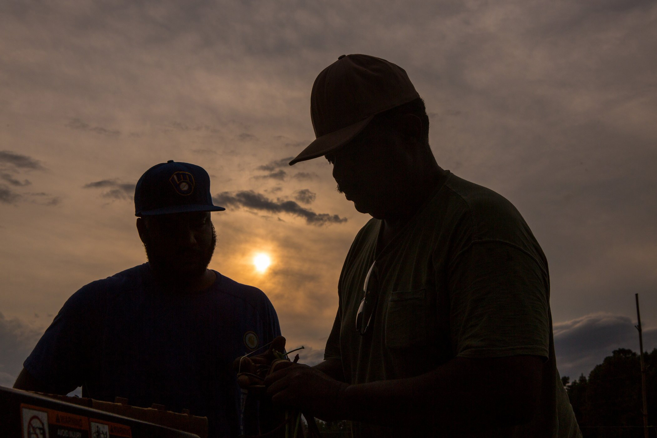 Two men silhouetted against a sunset, engaged in a discussion while handling equipment outdoors. The scene captures a moment of collaboration and connection in a natural setting.