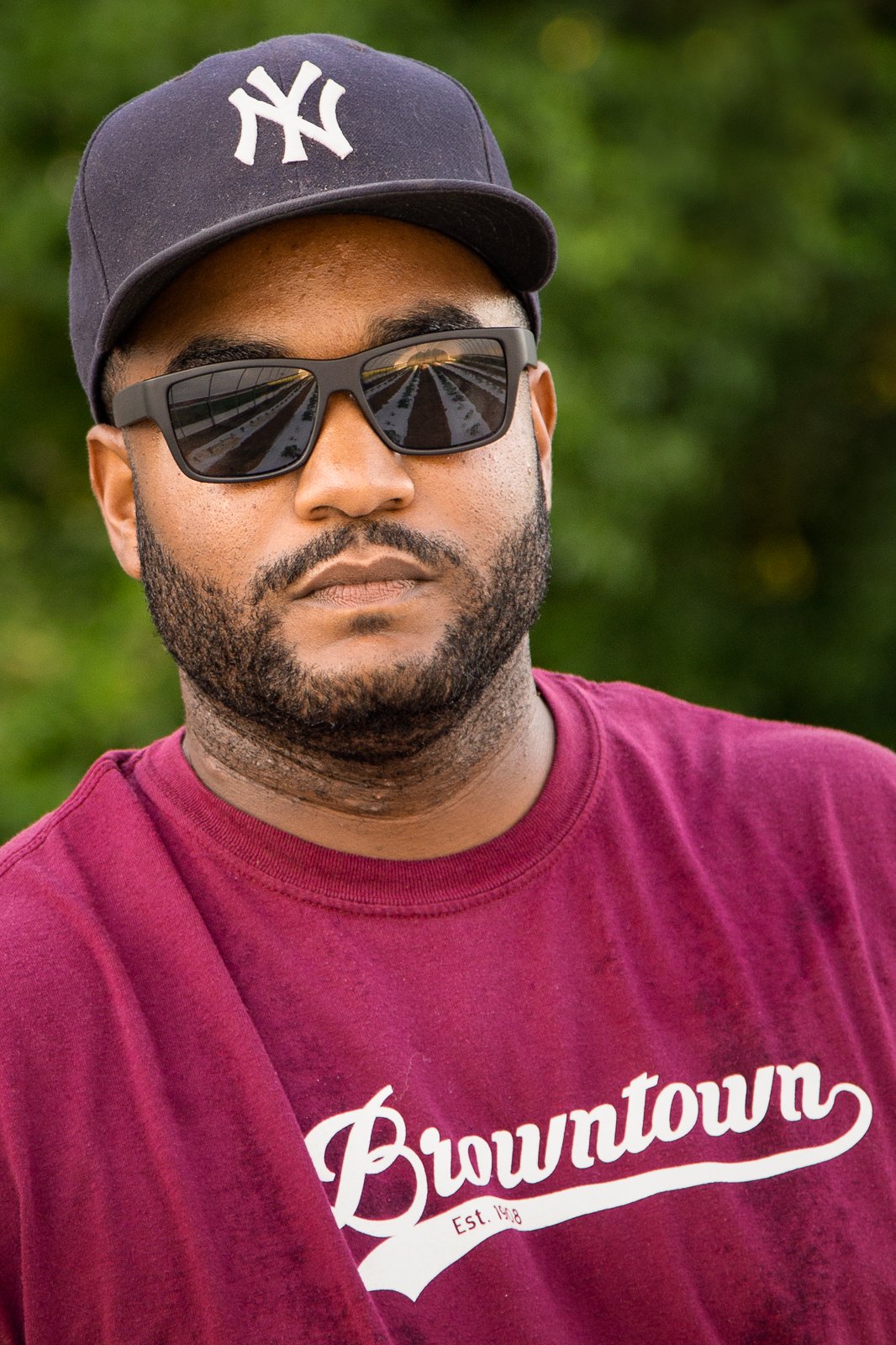 Man wearing sunglasses and a New York Yankees cap, posing confidently in a maroon shirt with 