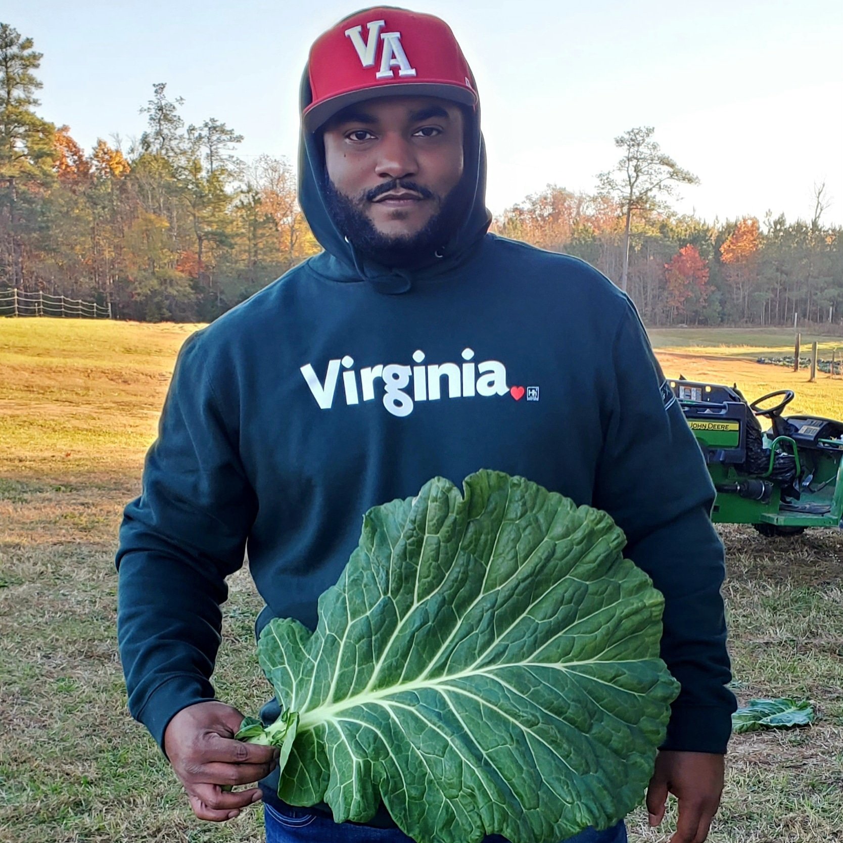 Man in a Virginia sweatshirt holding a large collard green leaf in a rural setting with a tractor in the background, showcasing local agriculture and farming culture.