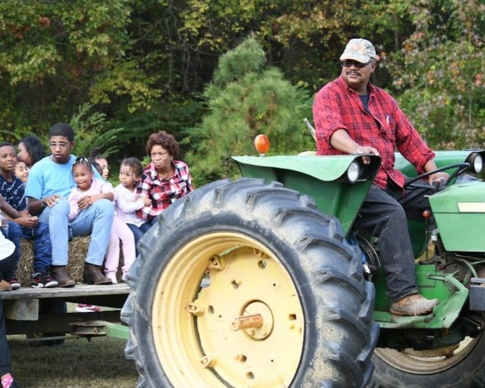 Herbert+Brown+-+Hay+Ride A man driving a green tractor while transporting a group of children and adults on a hayride, surrounded by trees in a rural setting. The scene captures a family-friendly outdoor activity, showcasing community and togetherness.