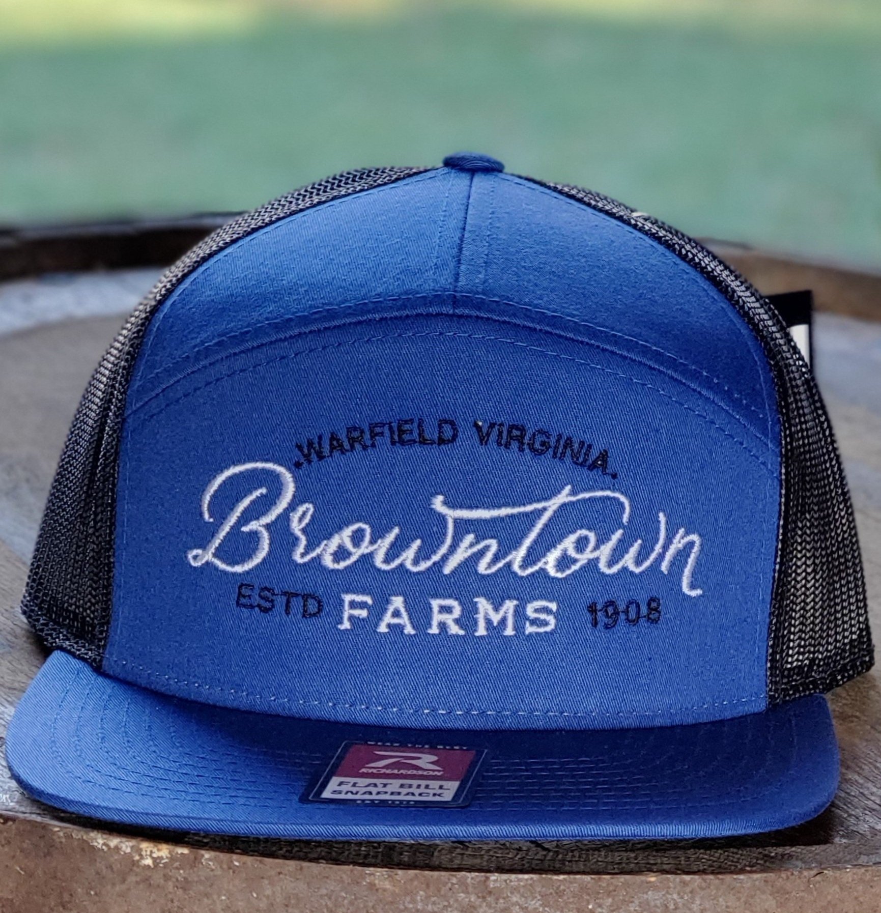 blue_crop Blue snapback hat featuring embroidered text "Warfield Virginia, Browntown Farms ESTD 1908," resting on a wooden surface with a blurred natural background.